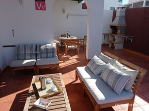 Une terrasse ensoleillée avec un banc en bois, un canapé confortable et une table sur laquelle sont disposés un plateau avec du café et des tasses.