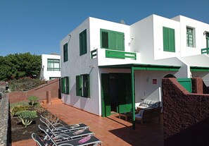 A modern building with white walls and green shutters, featuring a patio with sun loungers and a small seating area.
