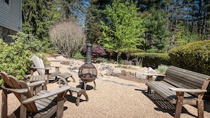 An outdoor seating area with wooden chairs and a bench, a metal fire pit, and a stone wall in the background. - Spacious & Gracious 3 Bedrooms, 2 Full Baths Ranch Living in central location!!! (Andover)