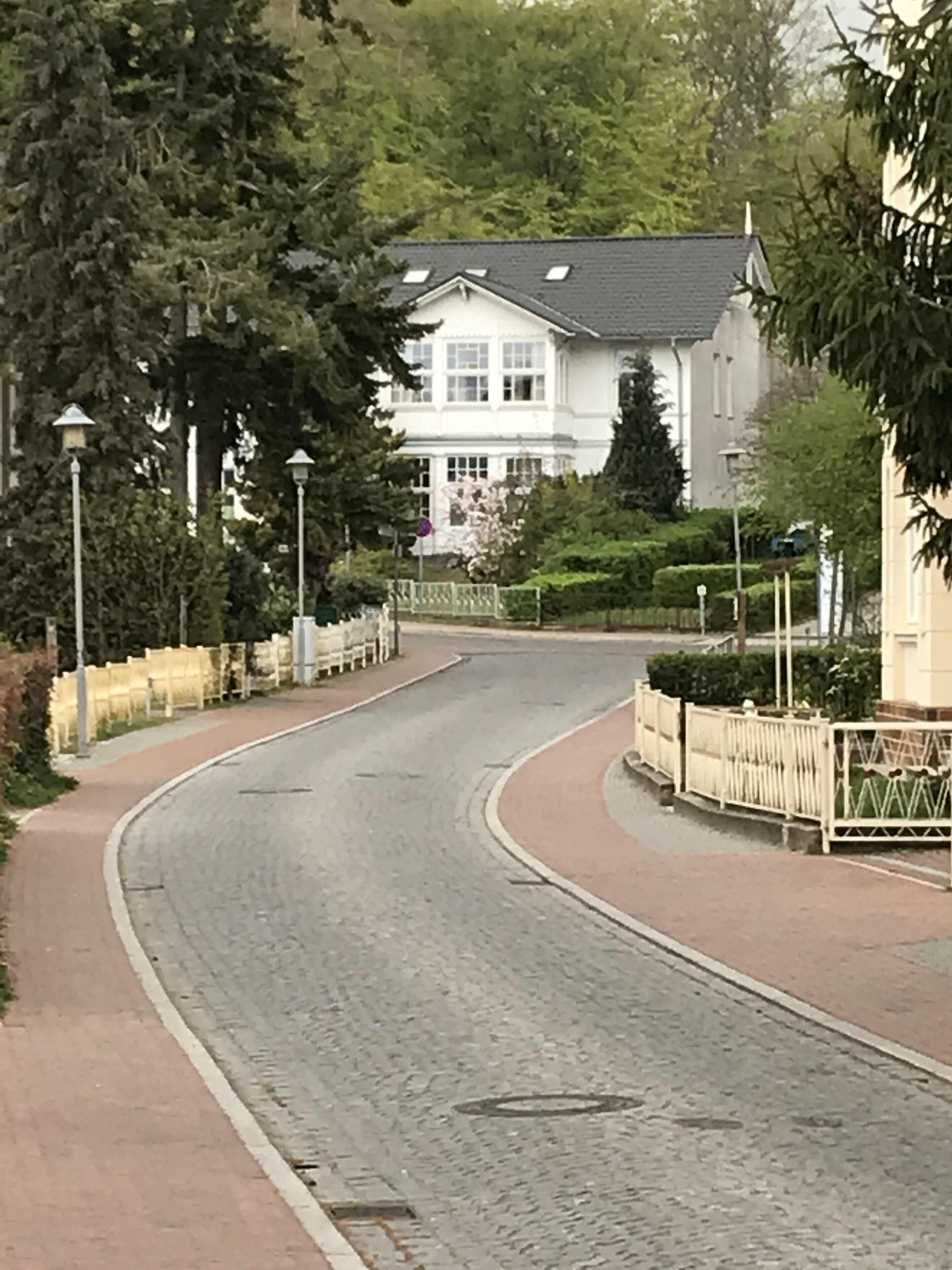 Une rue pavée et sinueuse, bordée d’arbres, et un bâtiment blanc surmonté d’un toit gris.