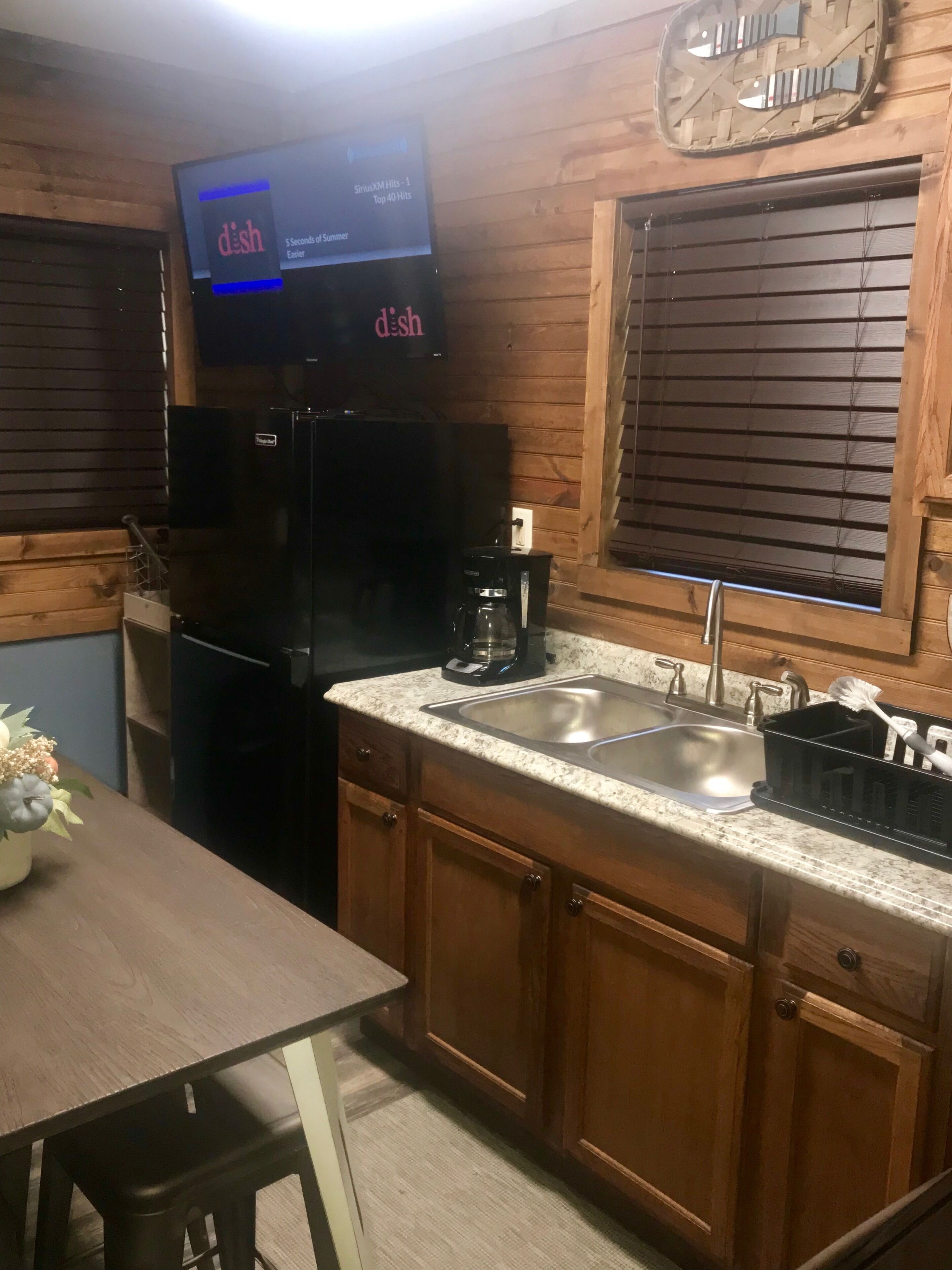 A compact kitchen with wooden cabinets, a black refrigerator, a coffee maker, and a TV displaying 'dish' on the screen.
