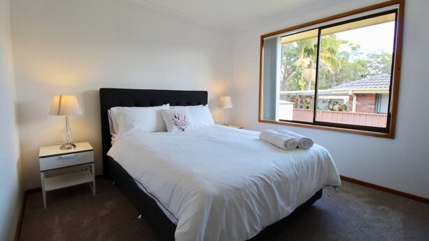 A neatly made bed with white linens, a black headboard, and a window with a view of a residential area. - Coastal Getaway by Book Jervis Bay - Free Wifi, linen & Games Room (Huskisson)