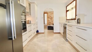 A modern kitchen with white cabinetry, stainless steel appliances, and a tiled floor. - Coastal Getaway by Book Jervis Bay - Free Wifi, linen & Games Room (Huskisson)