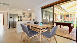A modern kitchen with stainless steel appliances, a dining area with a wooden table and grey chairs, and a pool table in an adjacent room. - Coastal Getaway by Book Jervis Bay - Free Wifi, linen & Games Room (Huskisson)