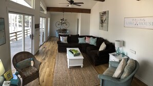 A spacious living room with a dark brown sofa, a white coffee table, and a wicker chair. A bicycle is mounted on the wall, and there is a sign that reads "The Reeves Family Lake House". - The Hideaway- Cozy Home in the Woods (Hot Springs)