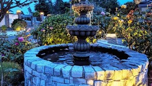 A stone fountain with multiple tiers, surrounded by a circular stone wall and various flowering plants. - Sparkling Clean, Private & Quiet! Amazing Ocean Views [6] (Brookings)