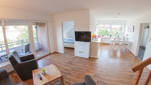Uma sala de estar moderna com uma televisão de tela plana, uma área de jantar com mesa e cadeiras, e grandes janelas com vista para o exterior.