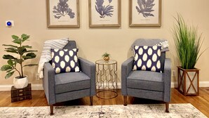 Deux fauteuils ornés de coussins à motifs, une petite table ronde sur laquelle est posée une plante, et trois reproductions botaniques encadrées accrochées au mur.