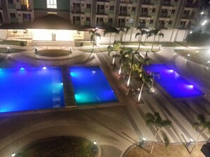 Vista nocturna d'un pati d'hotel amb dues piscines il·luminades, palmeres i un edifici al fons.