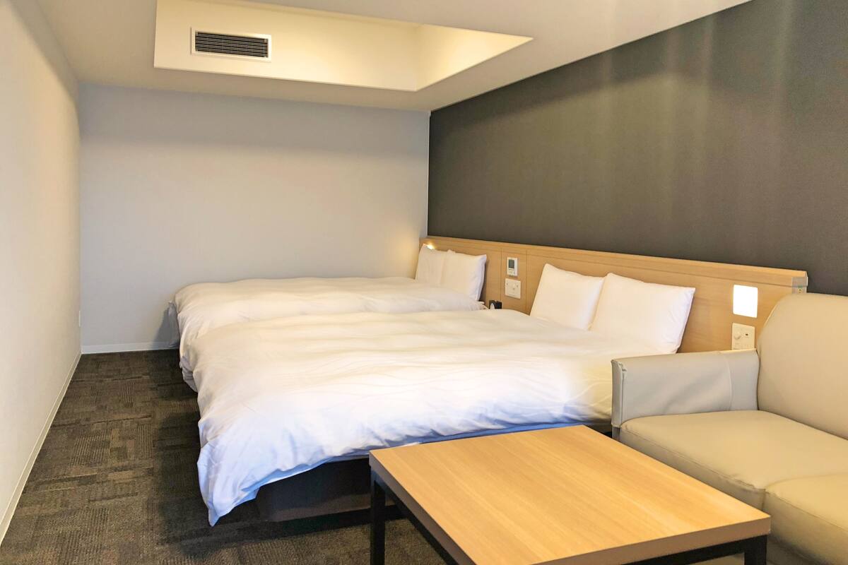 Habitación de hotel con una cama grande, una mesita de noche de madera, un sofá y una rejilla de ventilación en el techo.