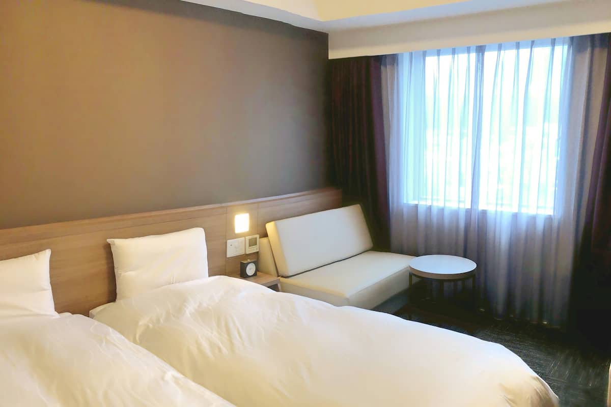 Habitación de hotel con dos camas grandes, un pequeño sofá, una mesa redonda y una ventana con cortinas.