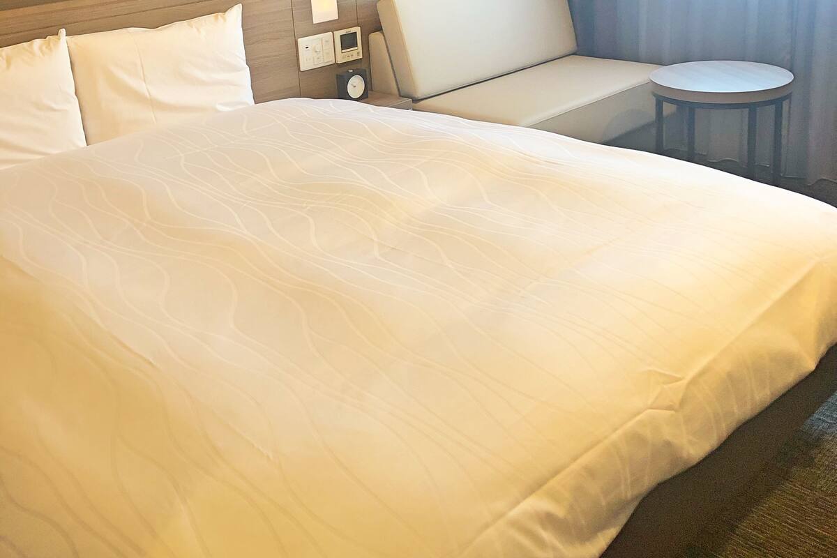 Habitación de hotel con una cama grande, un sofá, una mesita y un panel de control en la pared.