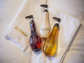 Toalha branca com a inscrição 'LA VISTA' bordada em dourado, colocada sobre uma cama com três produtos La Vista: sabonete corporal, condicionador e shampoo.
