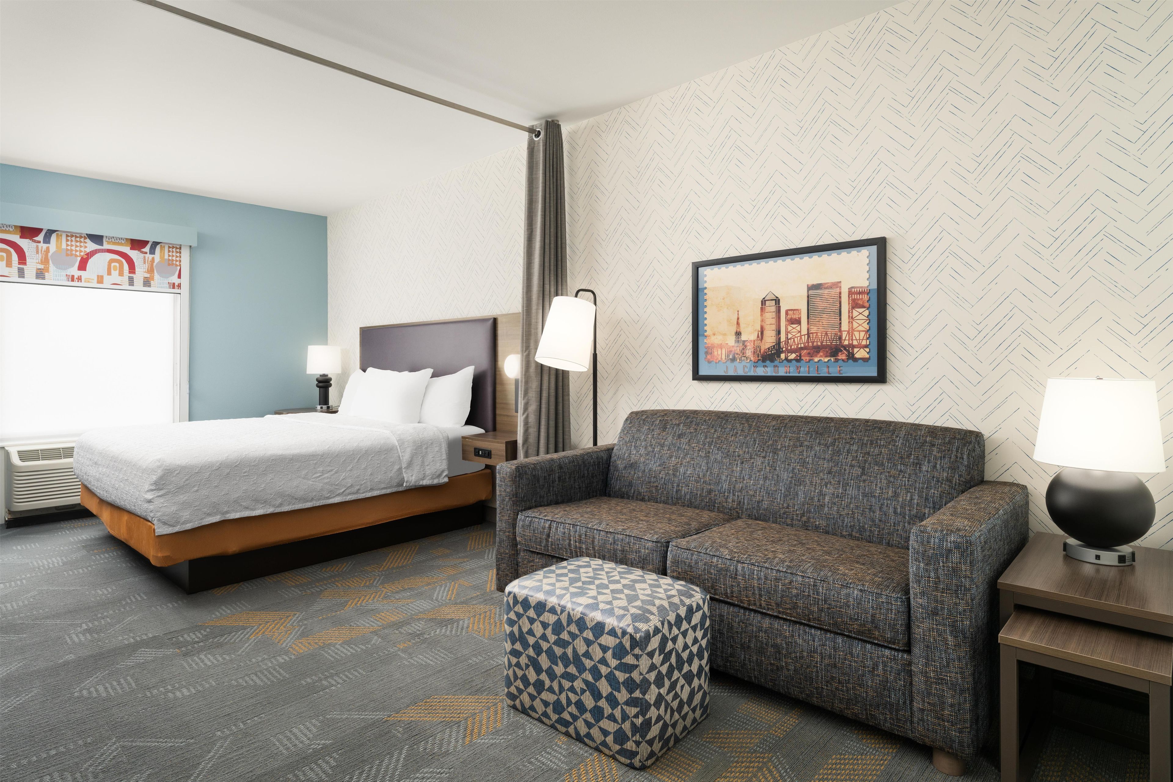 A hotel room with a bed, a sofa, a small ottoman, a nightstand, a lamp, and a framed picture on the wall.
