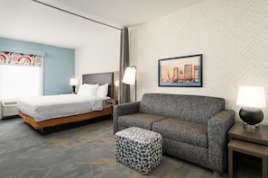 A hotel room with a bed, a sofa, a small ottoman, a nightstand, a lamp, and a framed picture on the wall. - Home2 Suites by Hilton Jacksonville Airport (Jacksonville)