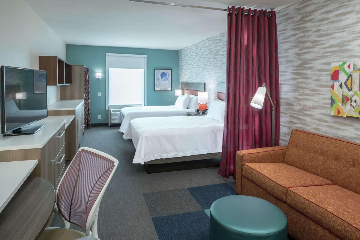 A hotel room with two beds, a desk with a TV, a sofa, and a window with curtains.