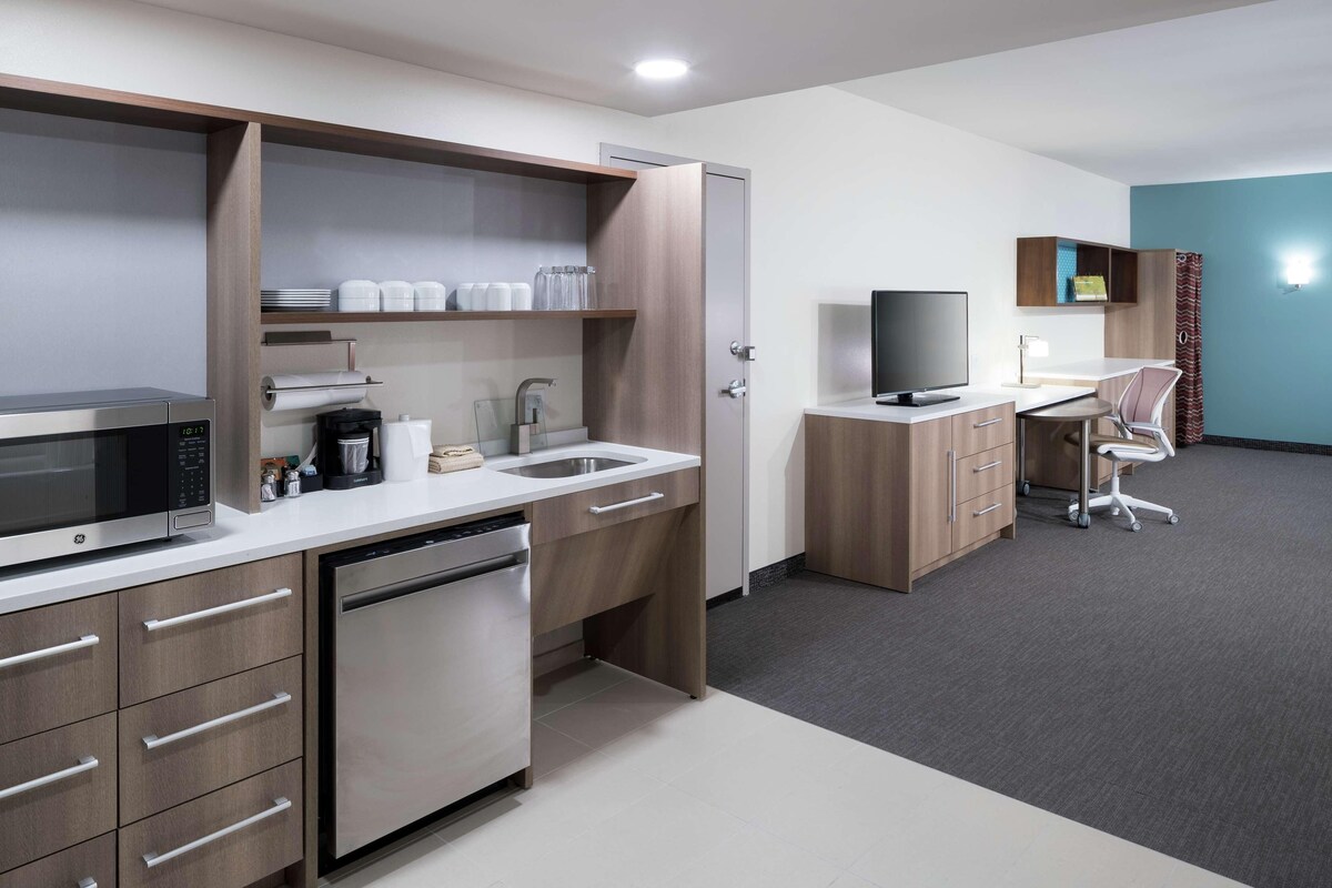 A modern kitchen with a microwave, coffee maker, and dishwasher. There is a wooden cabinet with a television and a desk with a chair.