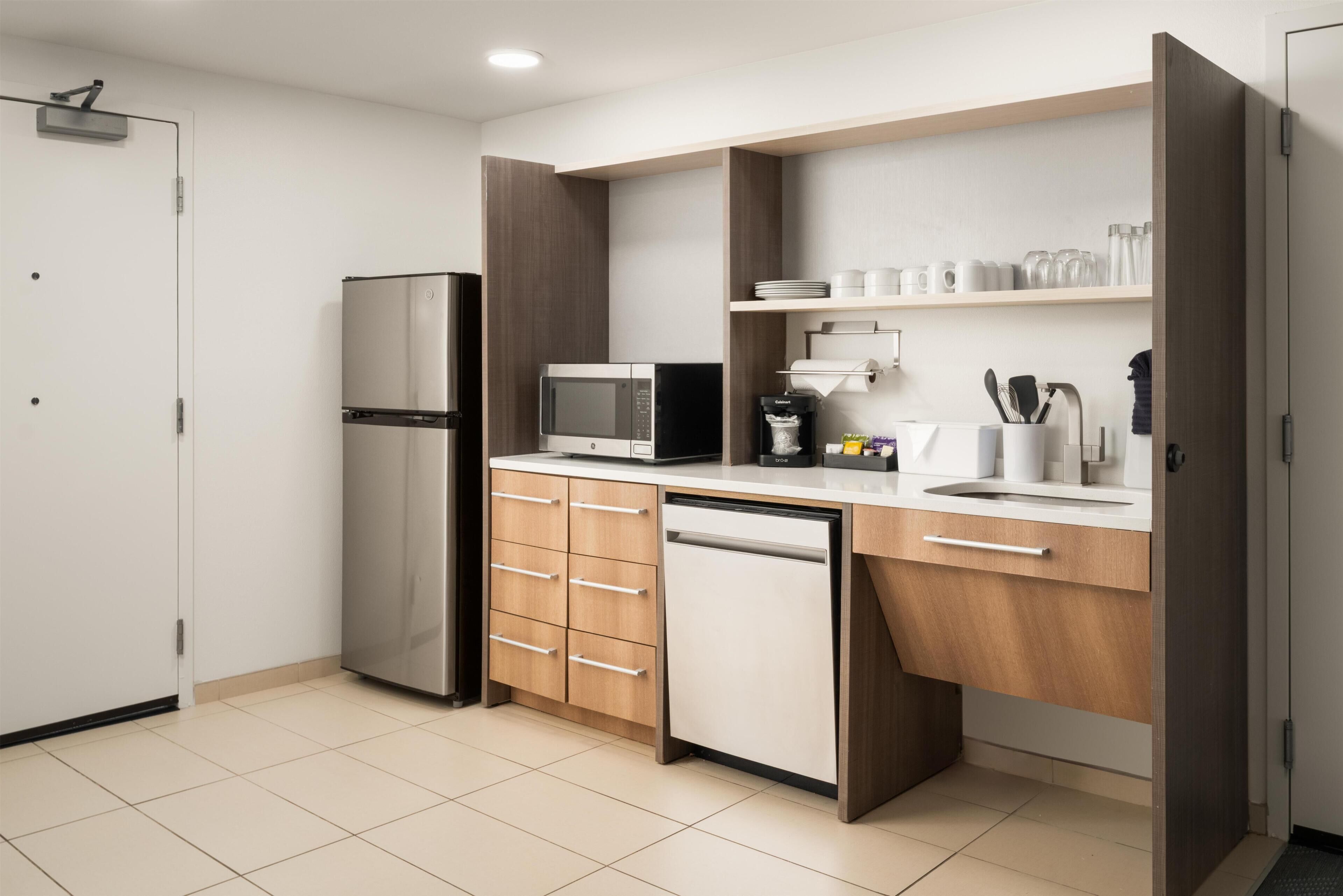A modern kitchen with a stainless steel refrigerator, a microwave, a dishwasher, wooden cabinets, and a white countertop.