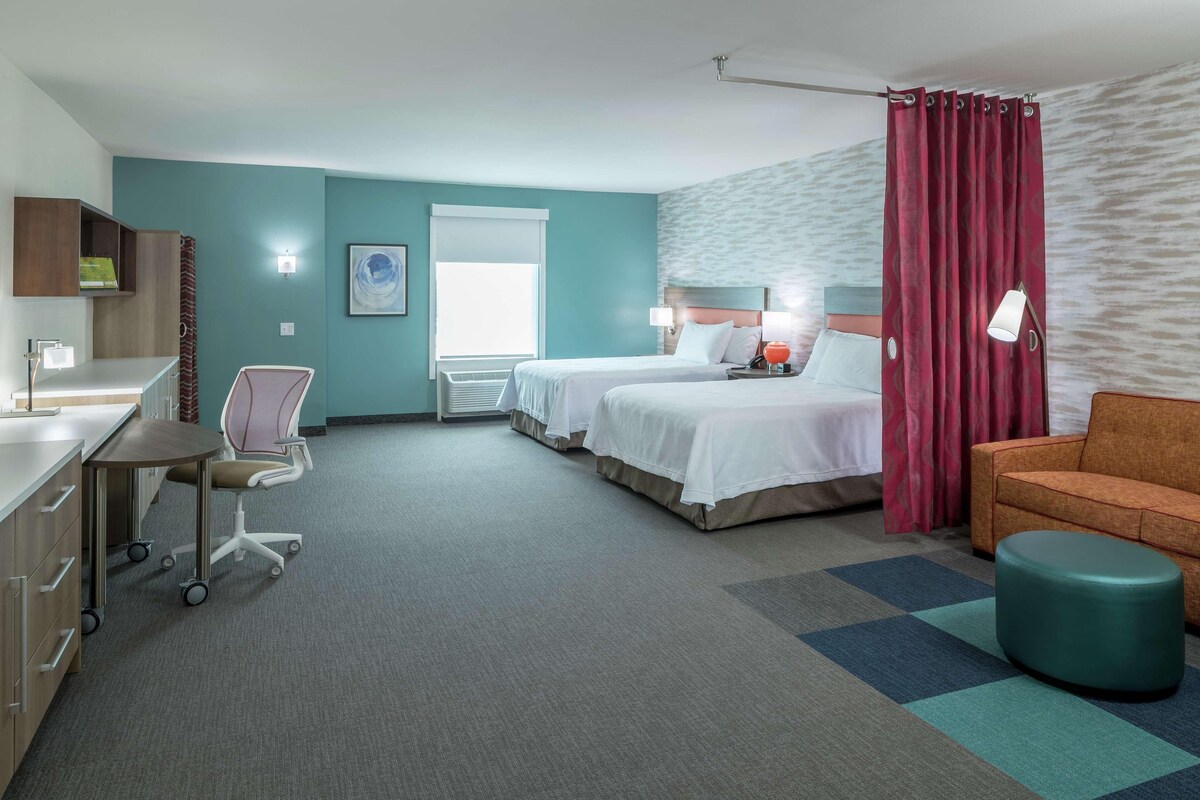 A hotel room with two beds, a desk, a chair, a sofa, and a small ottoman.