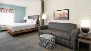 A hotel room with a bed, a sofa, an ottoman, a nightstand, a lamp, and a cityscape painting on the wall. - Home2 Suites by Hilton Jacksonville Airport (Jacksonville)