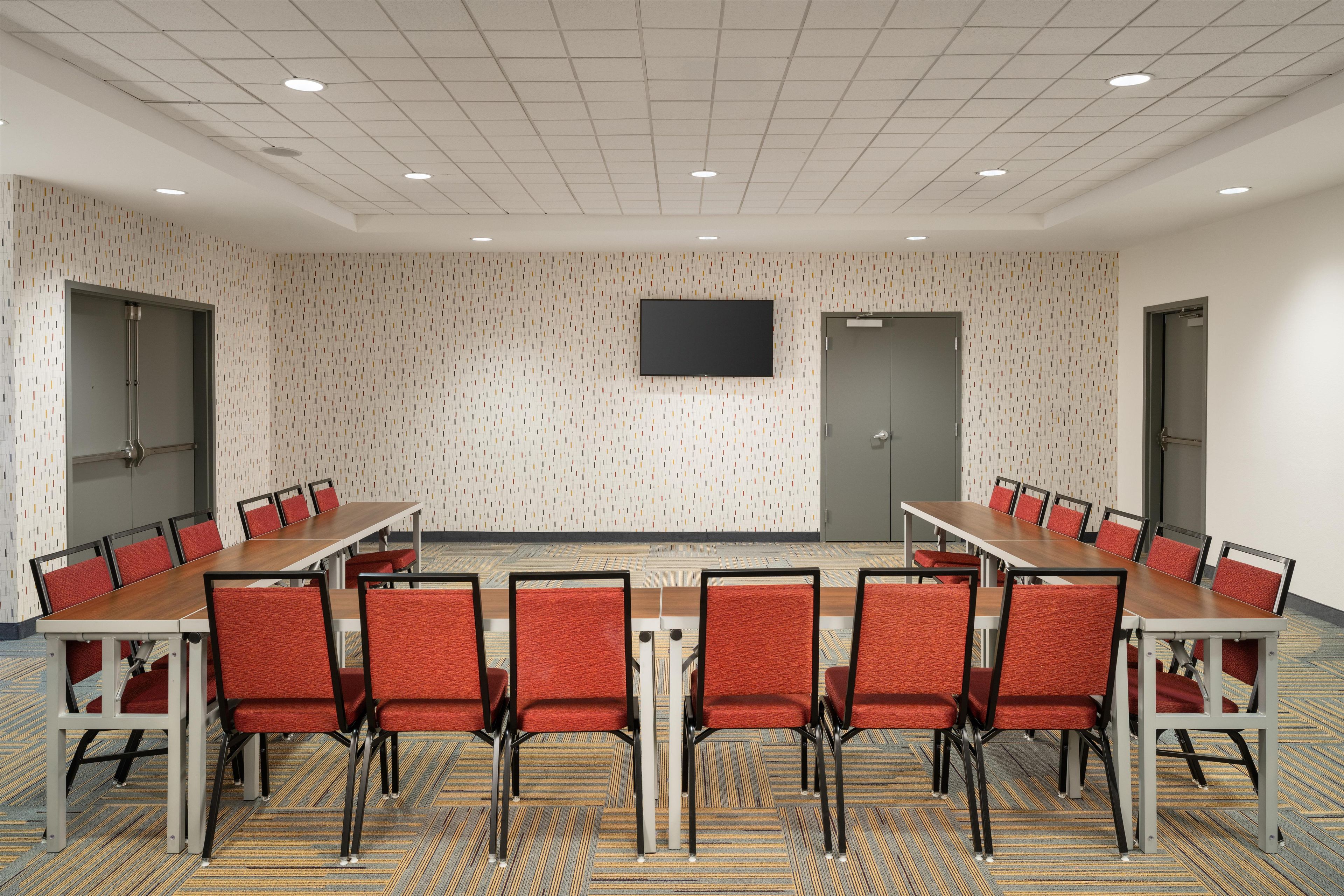 A conference room with a long table and red chairs, a flat-screen TV on the wall, and patterned carpet.