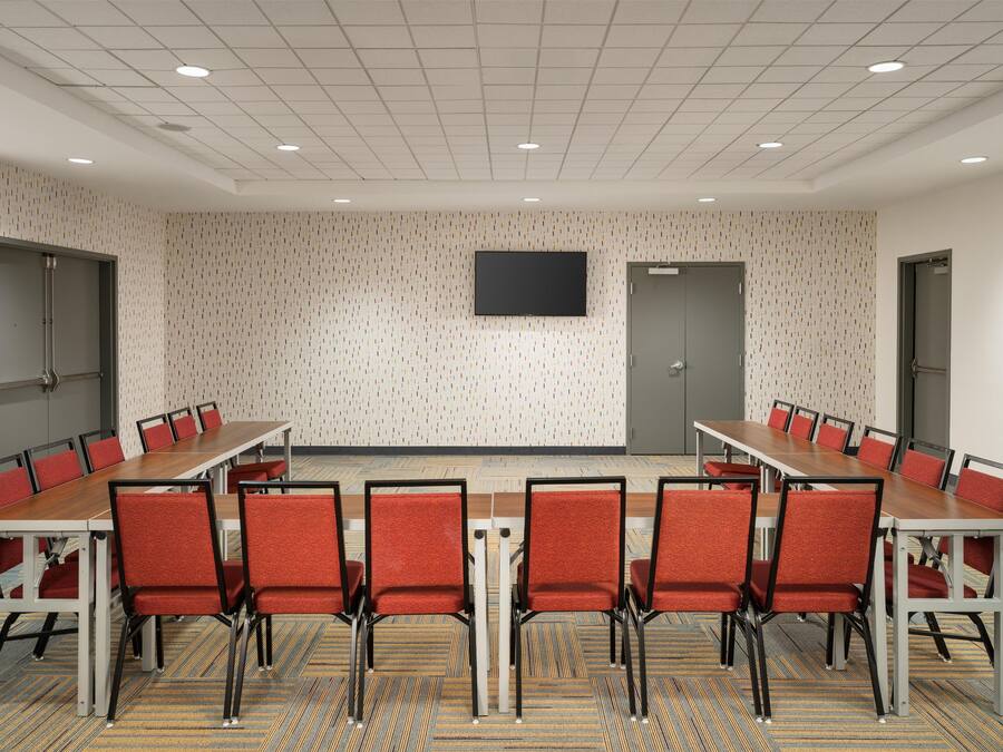 A conference room with a long table and red chairs, a flat-screen TV on the wall, and patterned carpet.