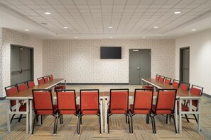 A conference room with a long table and red chairs, a flat-screen TV on the wall, and patterned carpet. - Home2 Suites by Hilton Jacksonville Airport (Jacksonville)