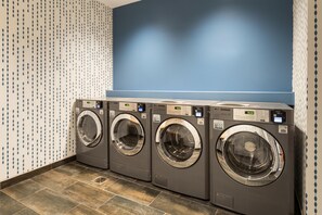 A row of four front-loading washing machines in a commercial setting. - Home2 Suites by Hilton Jacksonville Airport (Jacksonville)