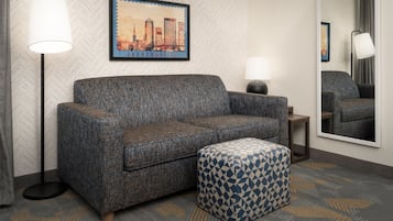 Ein Hotelzimmer mit Sofa, Hocker, Stehlampe, Beistelltisch und einem Bilderrahmen mit Stadtansicht.
