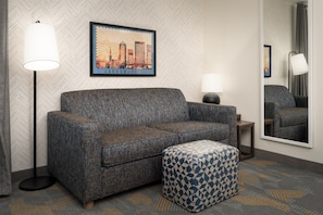 A hotel room with a sofa, an ottoman, a floor lamp, a side table, and a framed picture of a cityscape. - Home2 Suites by Hilton Jacksonville Airport (Jacksonville)