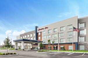 Un hotel Hampton Inn con diseño moderno, que cuenta con una entrada cubierta y un asta con la bandera estadounidense.