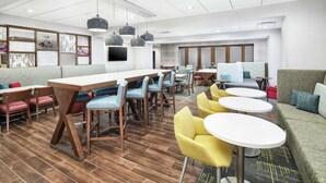 A modern hotel lobby with wooden flooring, multiple seating areas including a long communal table, individual chairs, and a sofa.