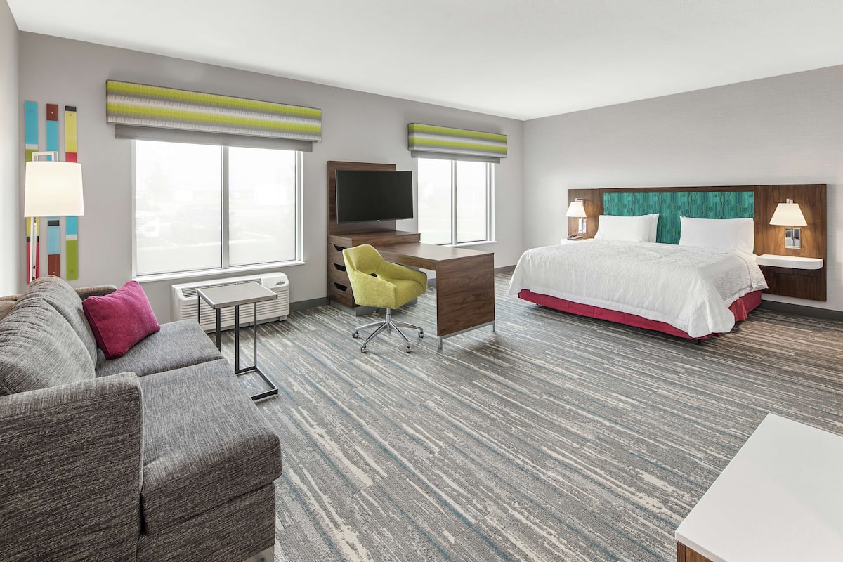A hotel room with a gray sofa, a yellow chair, a desk with a TV, a bed with white bedding, and a large window with blinds.