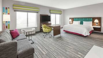 A hotel room with a gray sofa, a yellow chair, a desk with a TV, a bed with white bedding, and a large window with blinds.