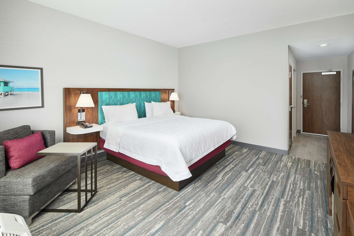 A hotel room with a bed, a sofa, a small table, a nightstand, and a wall-mounted sink.
