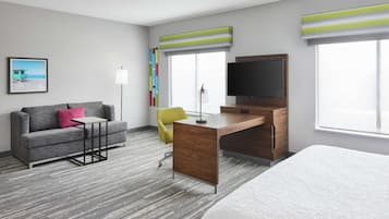 Habitación de hotel con sofá gris, una mesita, un escritorio con televisor y una cama.