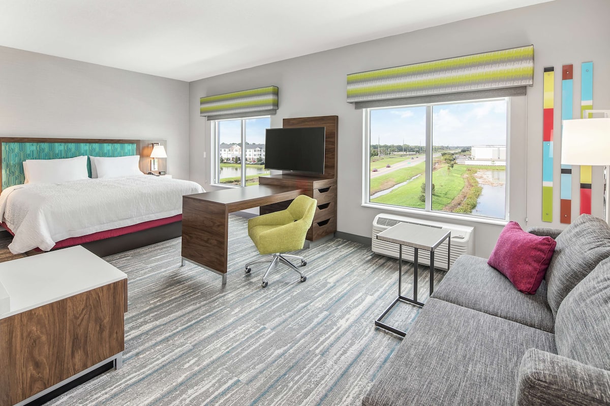 A hotel room with a bed, desk, and sofa, large windows with a view of a golf course, and a flat-screen TV.