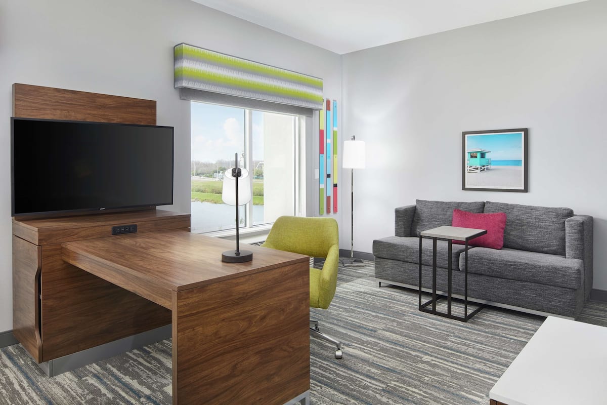A modern hotel room with a wooden desk, a flat-screen TV, a grey sofa, a small table, and a colorful window view.