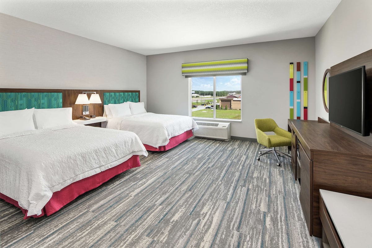 A hotel room with two beds, a desk, a chair, a TV, and a window with a view of a parking lot and buildings.