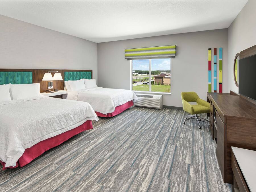 A hotel room with two beds, a desk, a chair, a TV, and a window with a view of a parking lot and buildings.