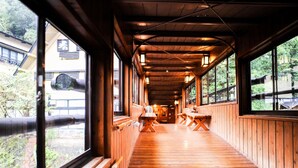 A wooden corridor with benches and tables, large windows, and a view of a building with a large pipe outside.