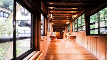 A wooden corridor with benches and tables, large windows, and a view of a building with a large pipe outside.