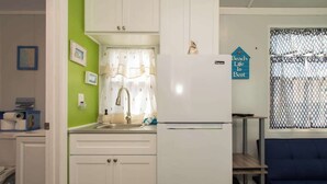 Une cuisine de taille réduite avec des meubles blancs, un évier et un réfrigérateur. Un panneau bleu est accroché au mur, et il y a une fenêtre avec un moustiquaire.