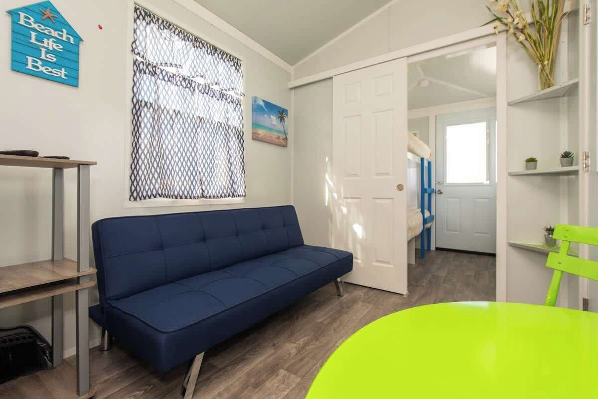 Una habitación con un sofá azul, una silla verde, una puerta blanca y una ventana con mosquitera.