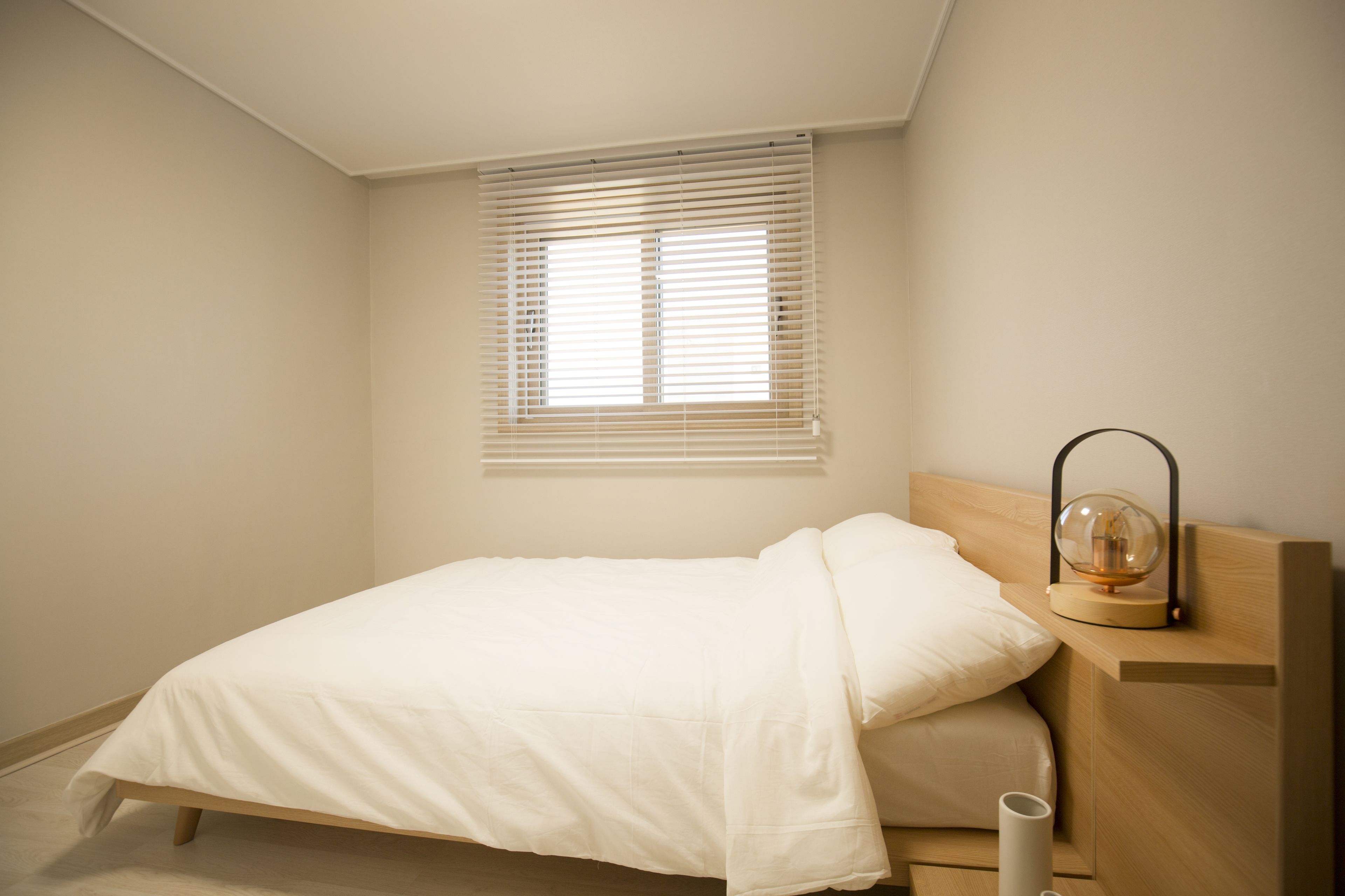 A minimalist bedroom with a single bed, a bedside table with a lamp, and a window with blinds.