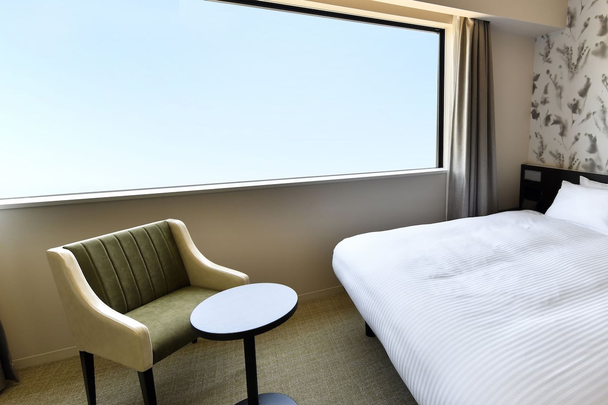 A hotel room with a bed, a chair, a small table, and a window.