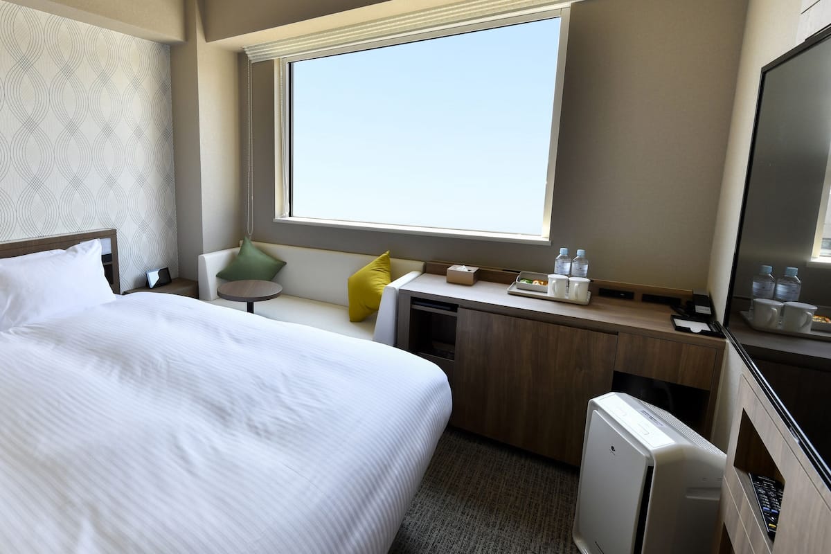 A hotel room with a large bed, a small desk, a chair, and a large window.