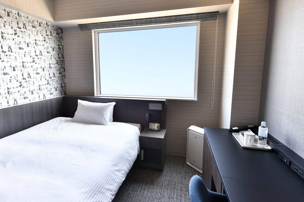 A hotel room with a bed, a desk, a chair, a small table with a bottle and a cup, and a window with a view.