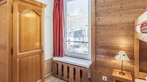 A room with a wooden wardrobe, a window with a view of buildings, a bench, and a bedside table with a lamp.