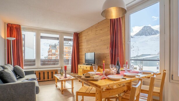 A cozy living room with a wooden table set for dining, a sofa, and a TV. Large windows offer a view of a snowy mountain.